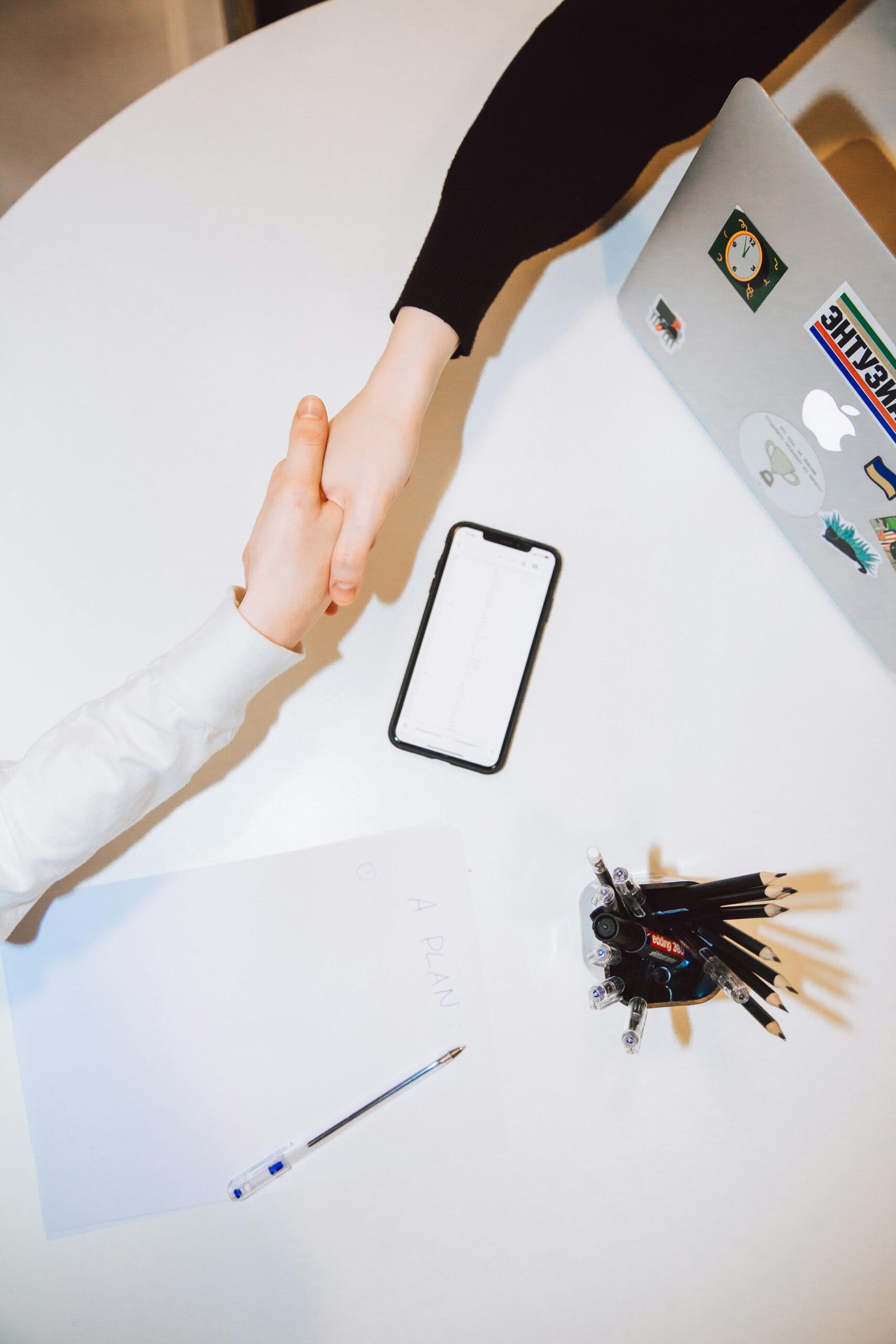pexels-photo-3746957-3746957 Two individuals shaking hands across a desk with a smartphone and laptop in a modern office setting.